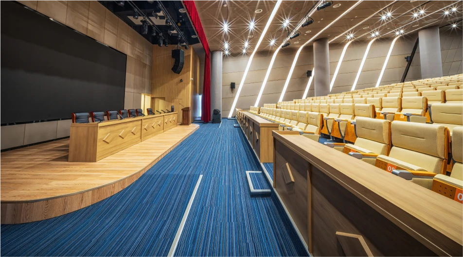 Spacious modern auditorium with stage, large screen, wooden paneling, blue carpet and rows of cushioned seats with translation devices