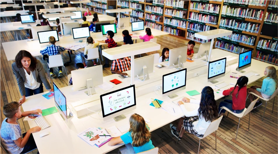 Modern library with children using desktop computers at shared white desks surrounded by bookshelves and guided by a teacher