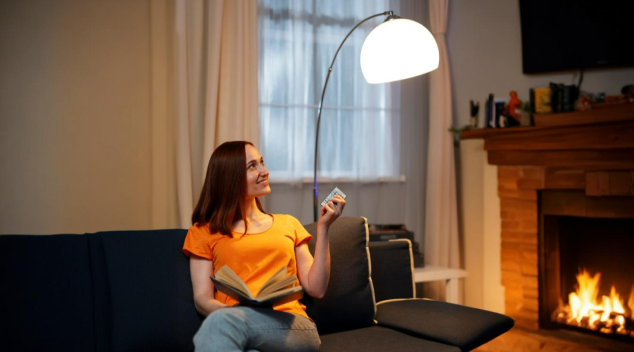 Woman adjusting smart lighting from couch in a cozy home environment with fireplace